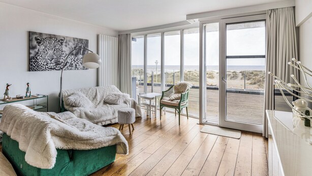 Une chambre moderne avec un canapé vert, un fauteuil blanc, une grande fenêtre donnant sur la plage et un tableau représentant un léopard.
