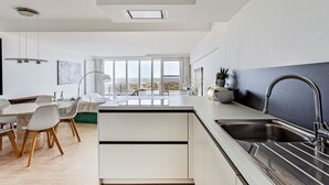 Une cuisine moderne dotée d’un îlot central, d’un coin repas avec une table ronde et des chaises, et de grandes fenêtres offrant une vue sur l’extérieur.