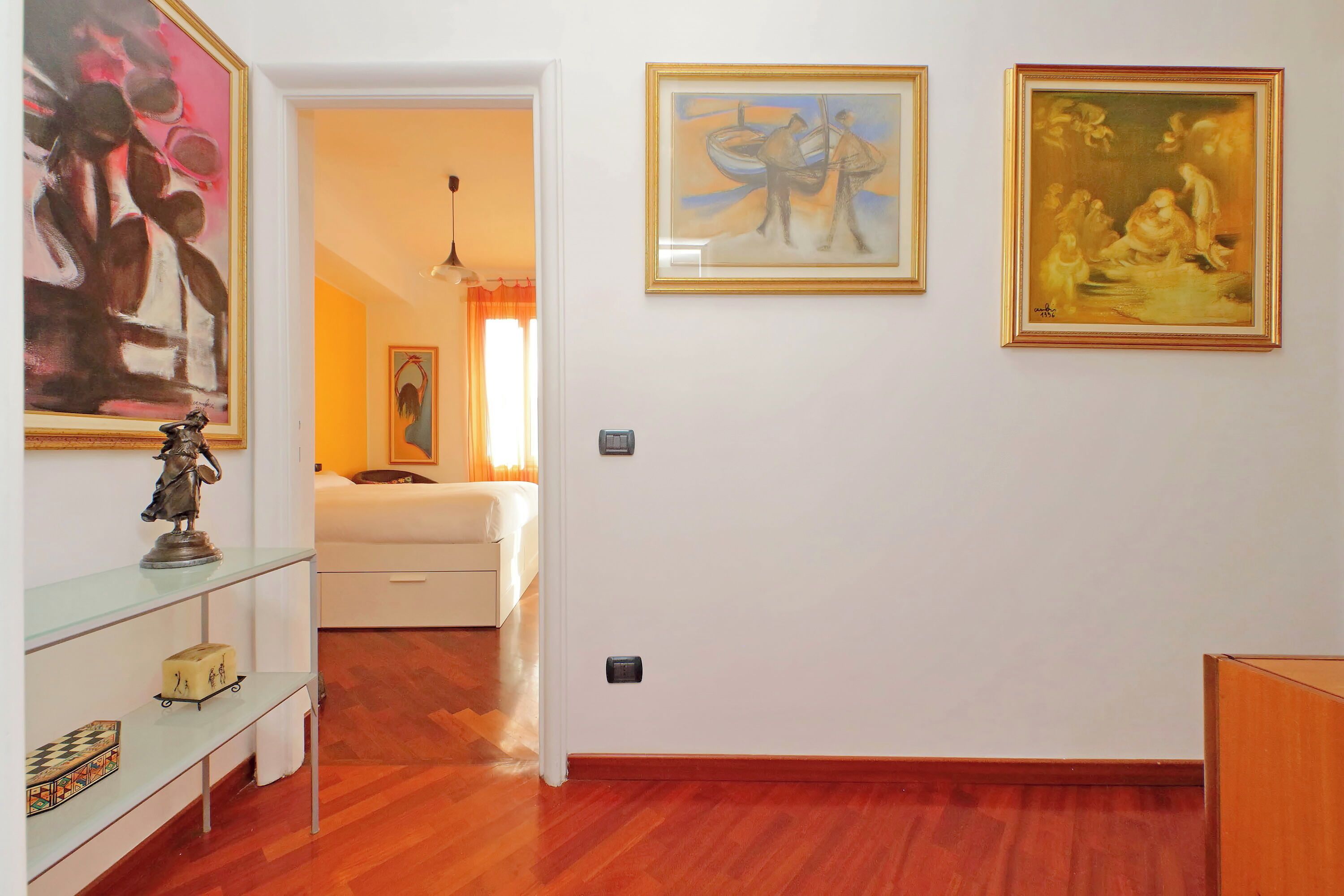 Une chambre d’hôtel avec un plancher en bois, un lit, une petite table sur laquelle se trouve une statue, et trois œuvres d’art encadrées accrochées au mur.