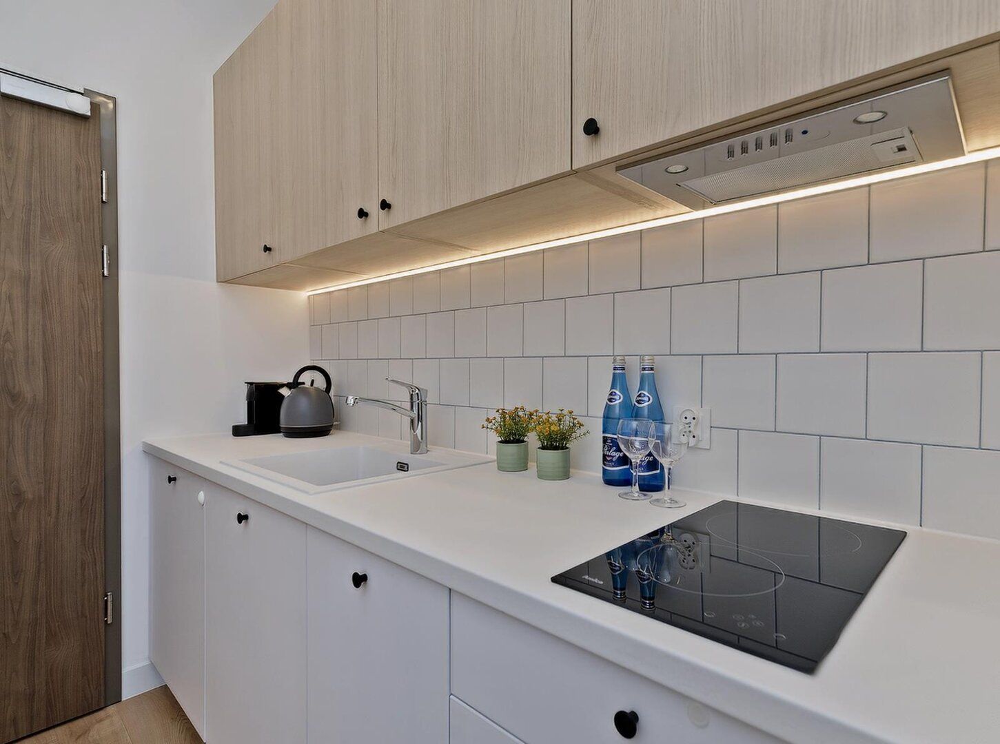 Una cocina moderna con armarios blancos, una placa de inducción negra, una tetera y dos plantas en macetas.