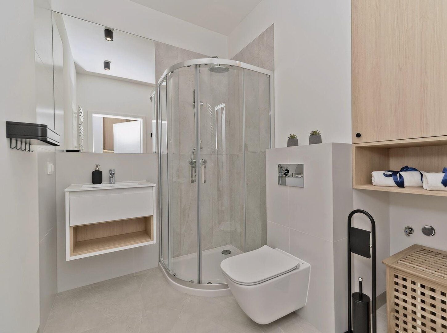 Un baño moderno con ducha de vidrio, inodoro blanco, lavamanos con espejo y estante de madera con toallas.