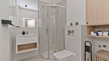 Un baño moderno con ducha de vidrio, inodoro blanco, lavamanos con espejo y estante de madera con toallas.