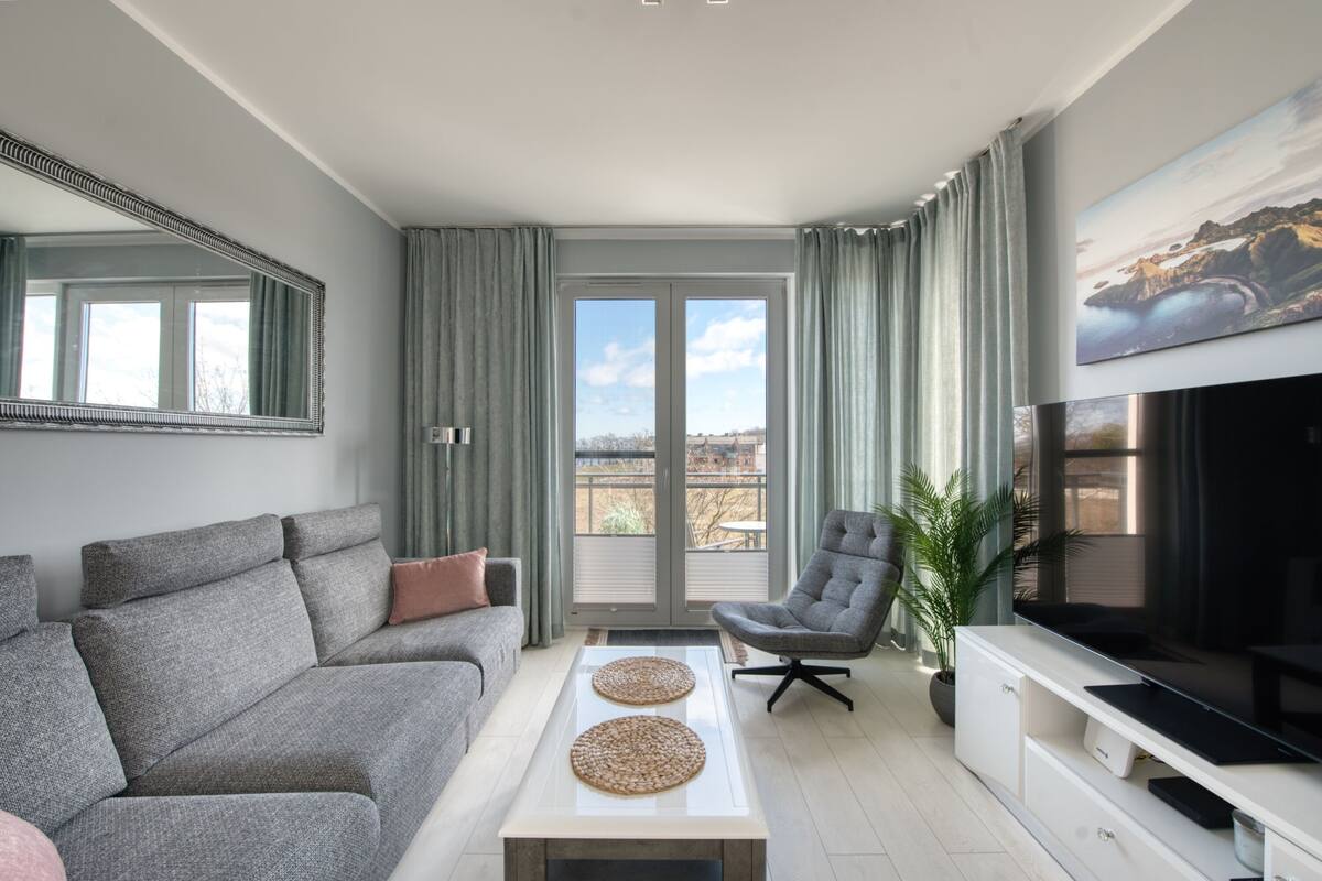 Un salon moderne avec un canapé gris, une table basse, un téléviseur et un balcon agrémenté de rideaux.