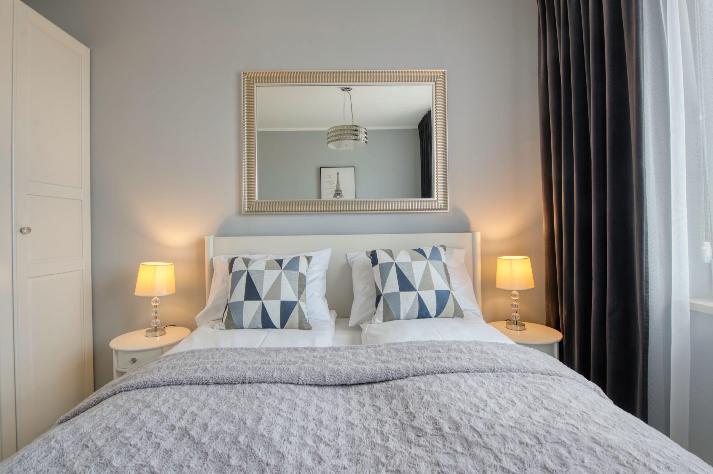 Een net opgemaakt bed met een kussen met dessin, een bedlampje en een spiegel erboven.