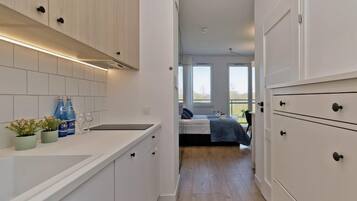 Un pasillo estrecho que da a un dormitorio con cama, ventana y vistas al exterior. Hay una zona de cocina con armarios, encimera y lavabo.