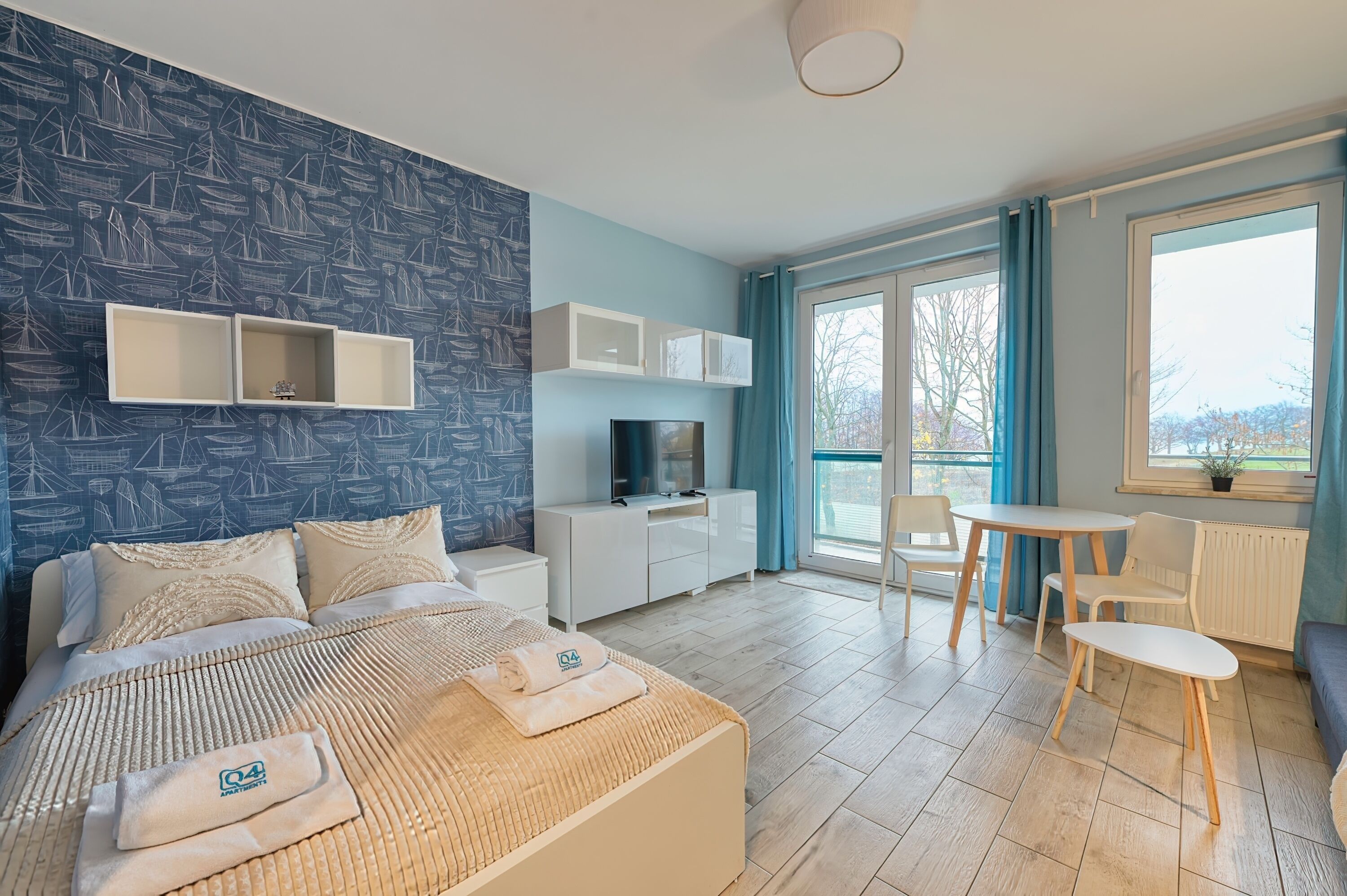 Habitación de hotel con cama, televisión, escritorio, sillas y balcón con vistas.
