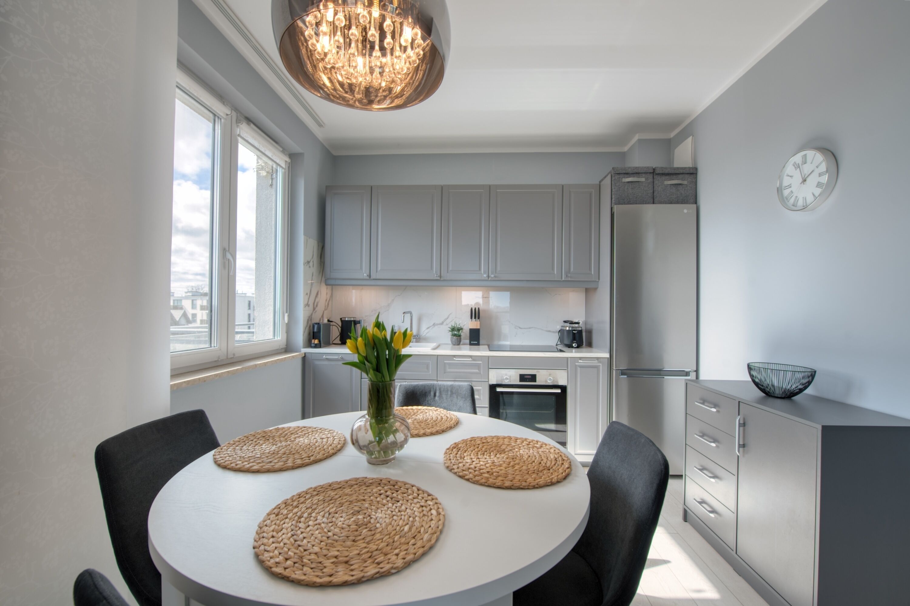 Een moderne keuken met grijze kastjes, een witte eettafel met geweven placematen, een kroonluchter en een klok aan de muur.