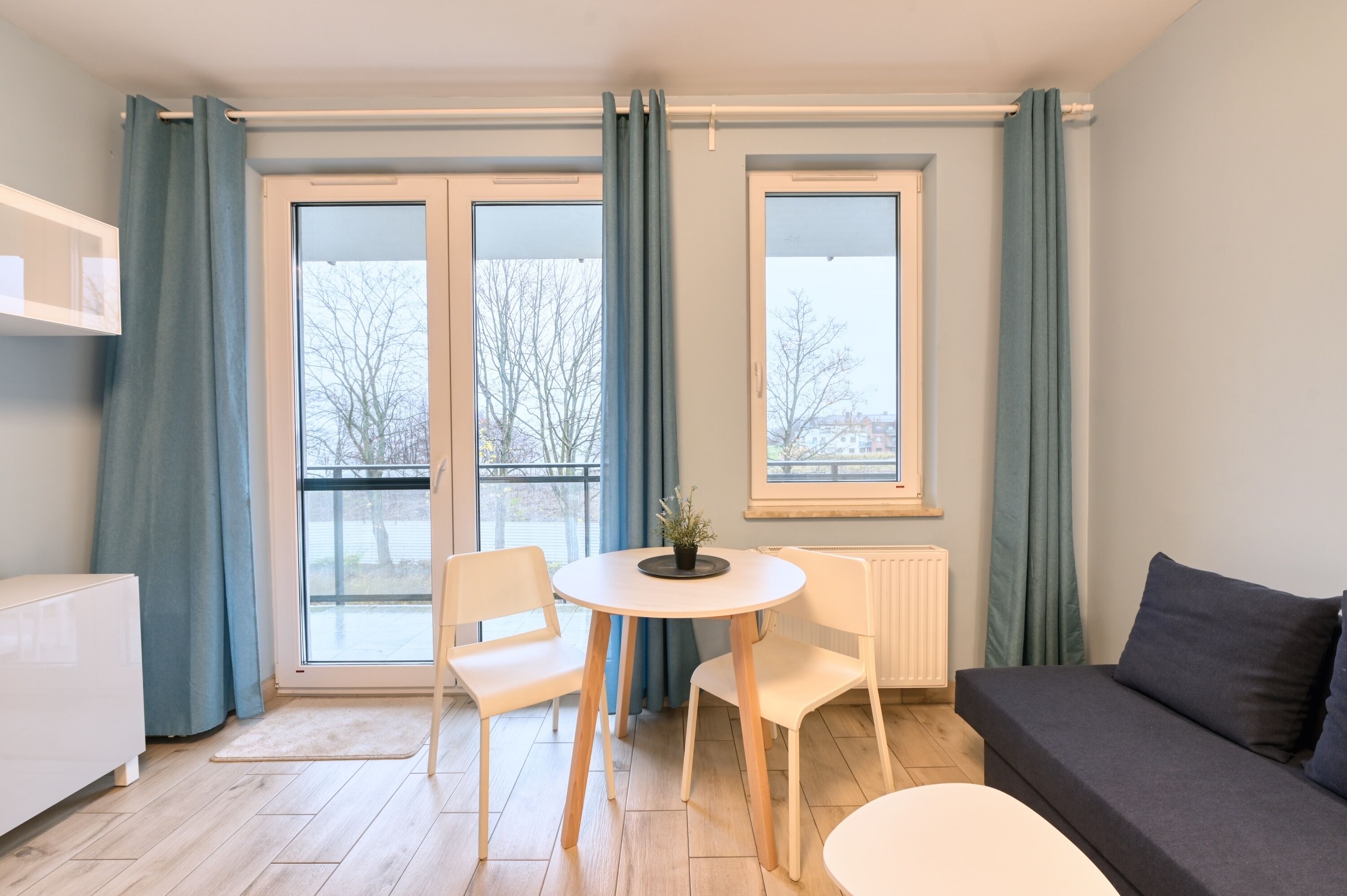 Una habitación moderna con sofá, una mesa de comedor con sillas, radiador y ventanas grandes con cortinas.