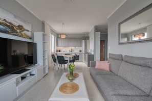 A modern living room with a flat-screen TV, a grey sofa, a coffee table, and a dining area with chairs and a table. - Apartamenty NEXO - Puck by Q4Apartments  (Puck)