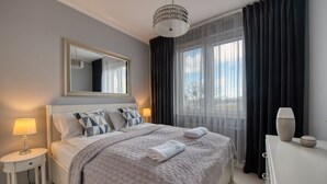 A neatly arranged bedroom with a bed, bedside tables, a mirror, and a window with curtains. - Apartamenty NEXO - Puck by Q4Apartments  (Puck)