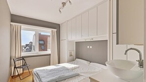 Ett sovrum med en säng, ett fönster med gardiner, ett element, en stol, ett sängbord och ett handfat med en spegel.