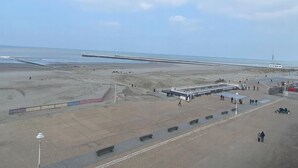 A beach with a long pier extending into the sea, a few people walking, and a small building near the water.