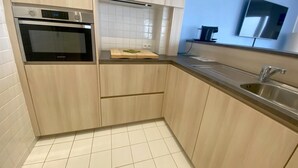A modern kitchen with light wood cabinets, a built-in oven, a stainless steel sink, and a wall-mounted clock.