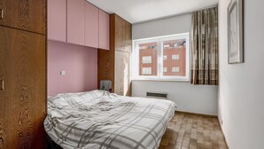 Ein Schlafzimmer mit Bett, Holzschrank, rosa Schränken, einem Fenster mit Blick auf ein Backsteingebäude und einer Heizung.