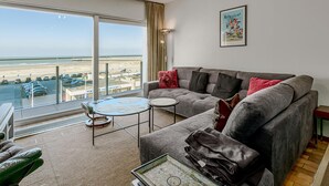 Ein modernes Wohnzimmer mit grauem Sofa, einem kleinen runden Tisch und Blick auf Strand und Meer durch große Fenster.