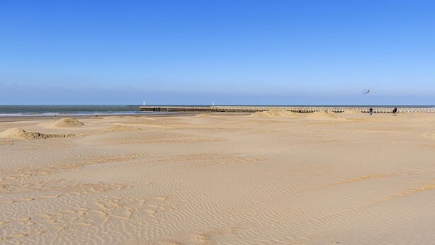 Ein Strand mit einer Seebrücke, die ins Wasser ragt, Sanddünen und einem klaren Himmel.