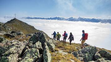 Eine Gruppe Wanderer auf einem felsigen Bergpfad mit einem Kreuz auf einem Berggipfel, umgeben von Wolken und fernen Bergen.