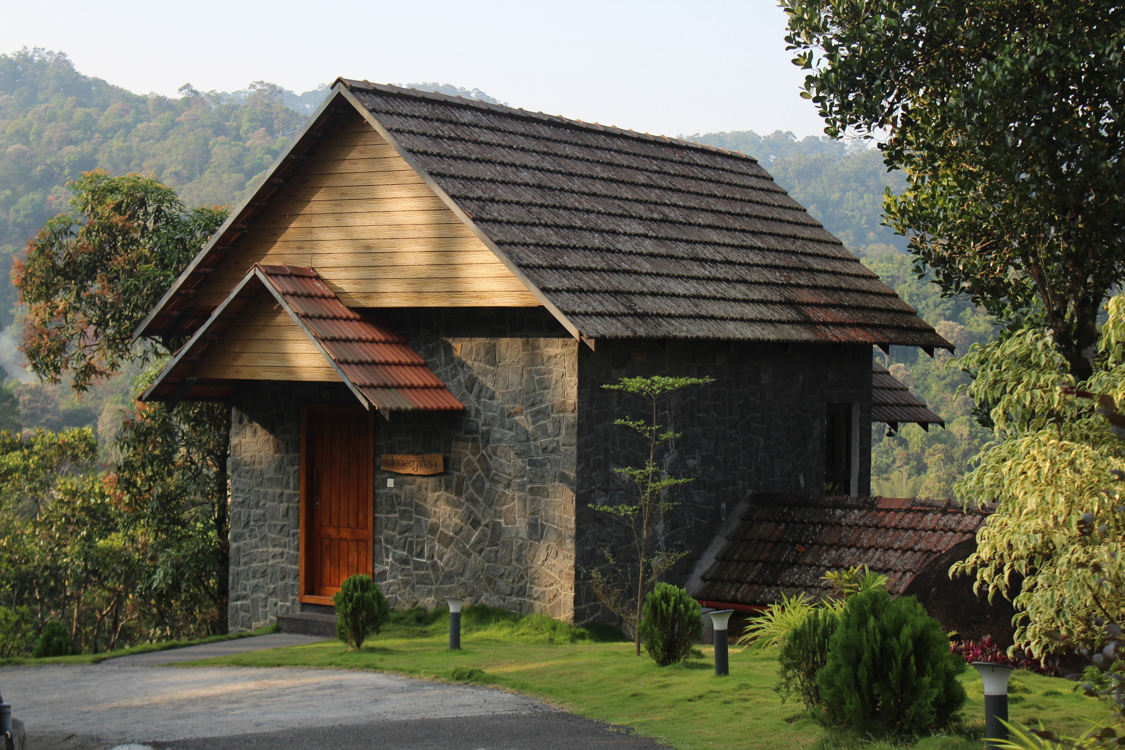 A stone building with a wooden facade and a sloped roof, surrounded by lush greenery and hills in the background.