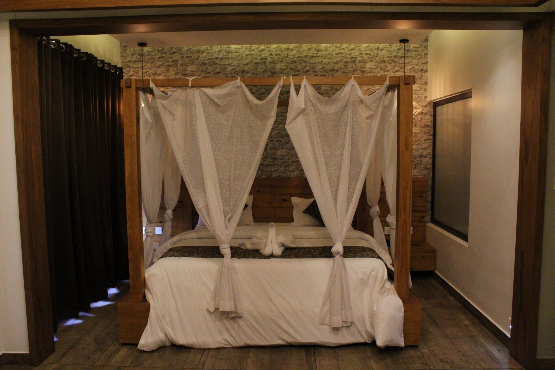 A four-poster canopy bed with a stone wall backdrop, wooden headboard, and a large window to the right.