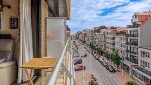 A balcony with a wooden table and chairs, overlooking a street with parked cars and buildings.