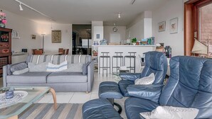 En moderne stue med en grå sofa, et glasbord og en blå lædersofa. I baggrunden er der et køkkenområde med en bar og barstole.