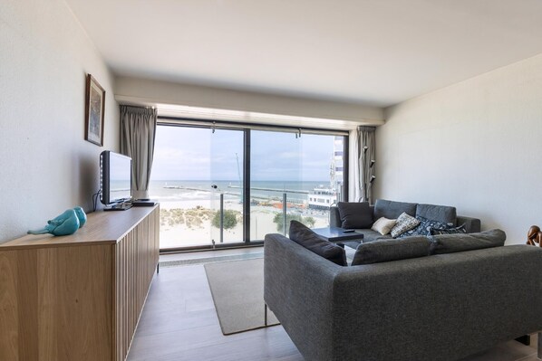 Een moderne woonkamer met een grijze bank, een houten bijzettafeltje en een groot raam met uitzicht op het strand en de skyline.