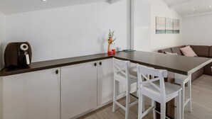 Una cocina moderna con armarios blancos, encimera oscura y una cafetera. Hay una silla blanca y un florero con flores sobre la encimera.