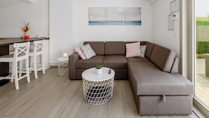 Una sala de estar moderna con un sofá seccional, una mesa de centro redonda con una planta, una zona de comedor blanca con sillas y un ventanal con vistas.