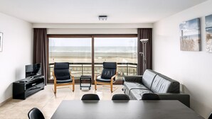 Moderni hotellihuone, jossa on suuri parveke, sohva, kaksi tuolia, televisio ja ruokapöytä.