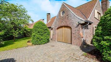 Een bakstenen huis met een houten deur, een verharde oprit en veel groen eromheen.