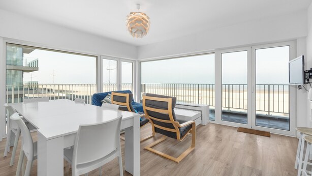 En rummelig stue med et stort hvidt bord, to stole, en moderne loftlampe og store vinduer med udsigt over stranden.