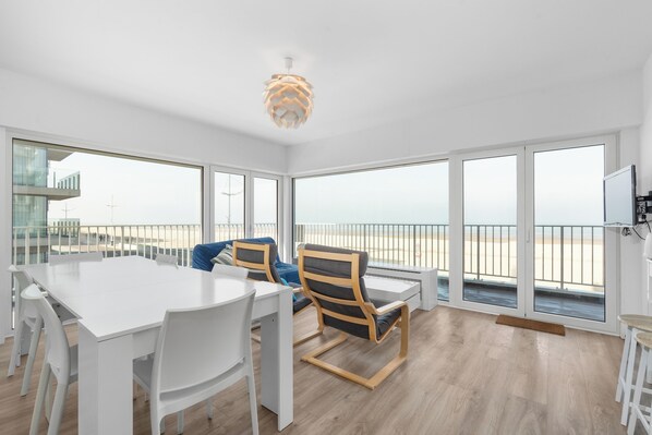 Amplia habitación con una gran mesa blanca, dos sillas, una lámpara de techo moderna y ventanas amplias que ofrecen vista a la playa.