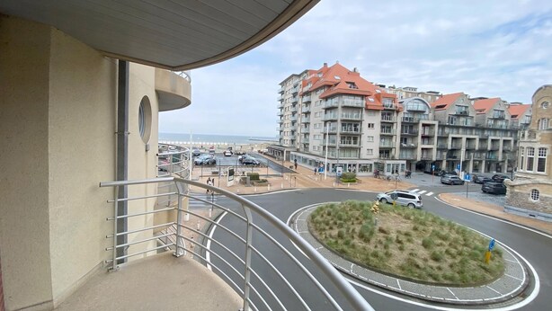 A balcony with a view of a roundabout, a parking area, and a building with multiple balconies and a red roof.