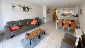 A modern living room with a grey sofa, a wooden coffee table, and a dining area with orange chairs.