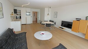 Uma sala de estar moderna com um sofá, uma mesa de centro redonda com uma tigela, uma área de jantar com cadeiras e uma mesa, uma cozinha com armários e eletrodomésticos, e uma TV sobre um suporte.