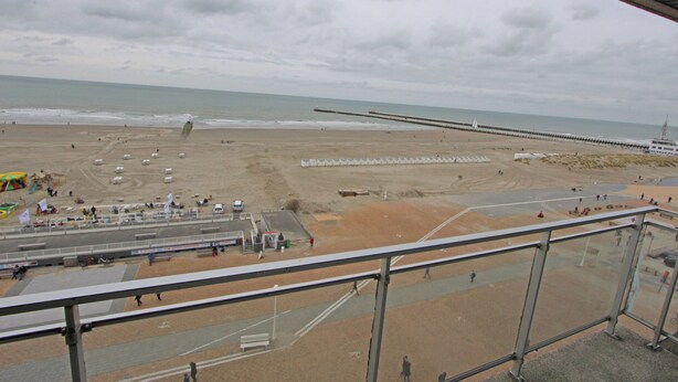 Uma praia com um longo pier que se estende sobre a água, uma área arenosa com pessoas e veículos, e um céu claro.