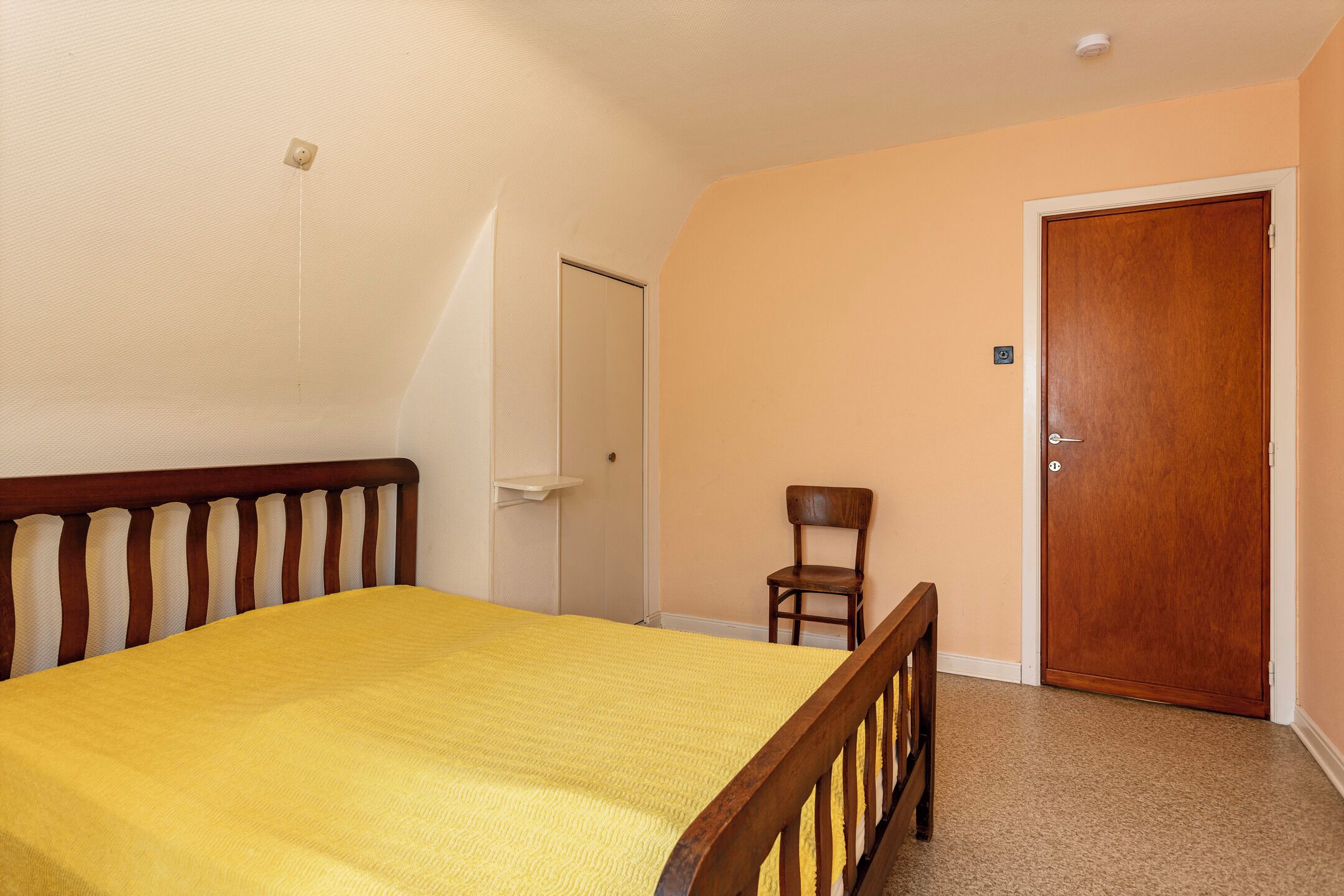 A bedroom with a bed, a chair, a door, and a light switch on the wall.