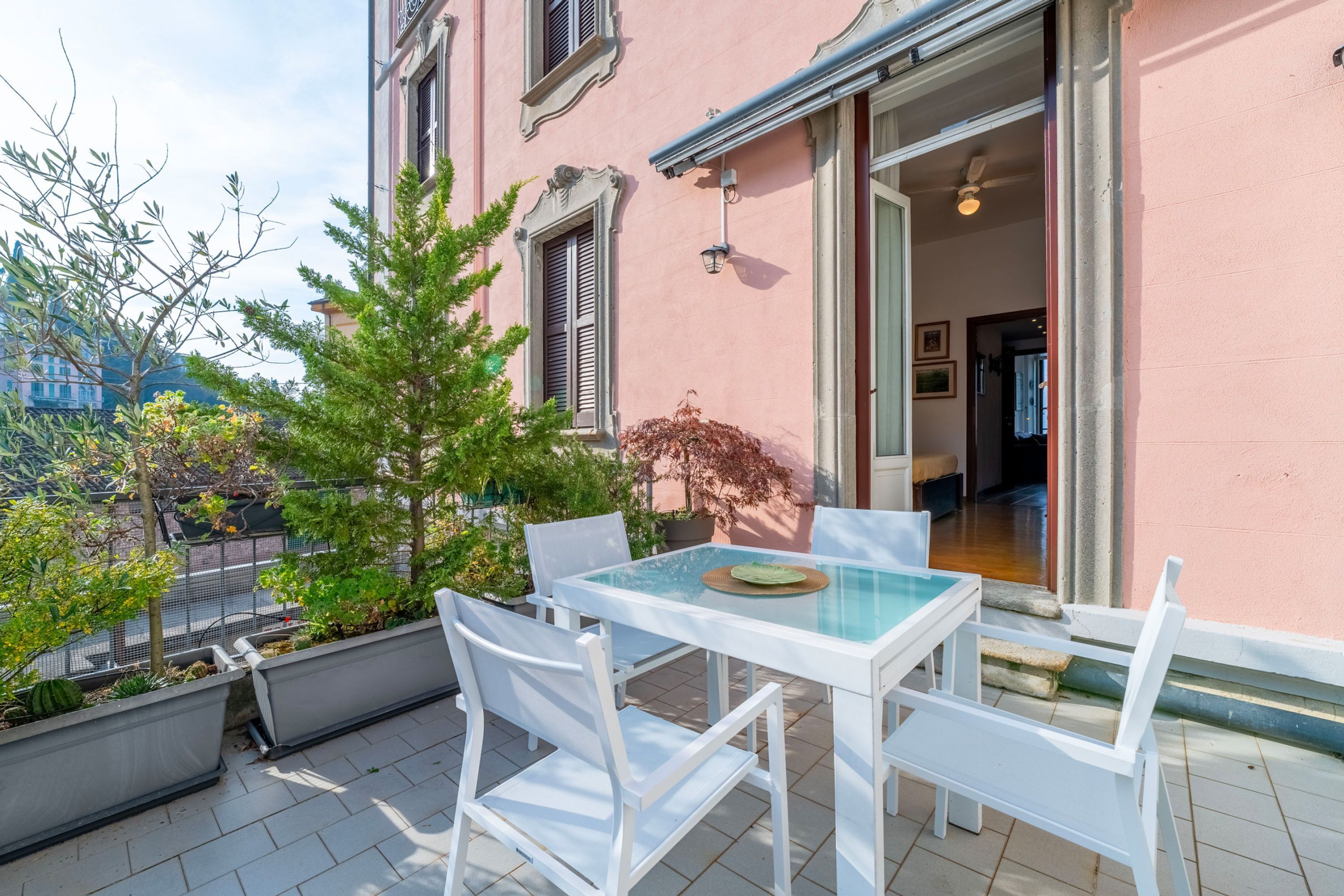 A balcony with a glass table and white chairs, overlooking a street with buildings and trees.