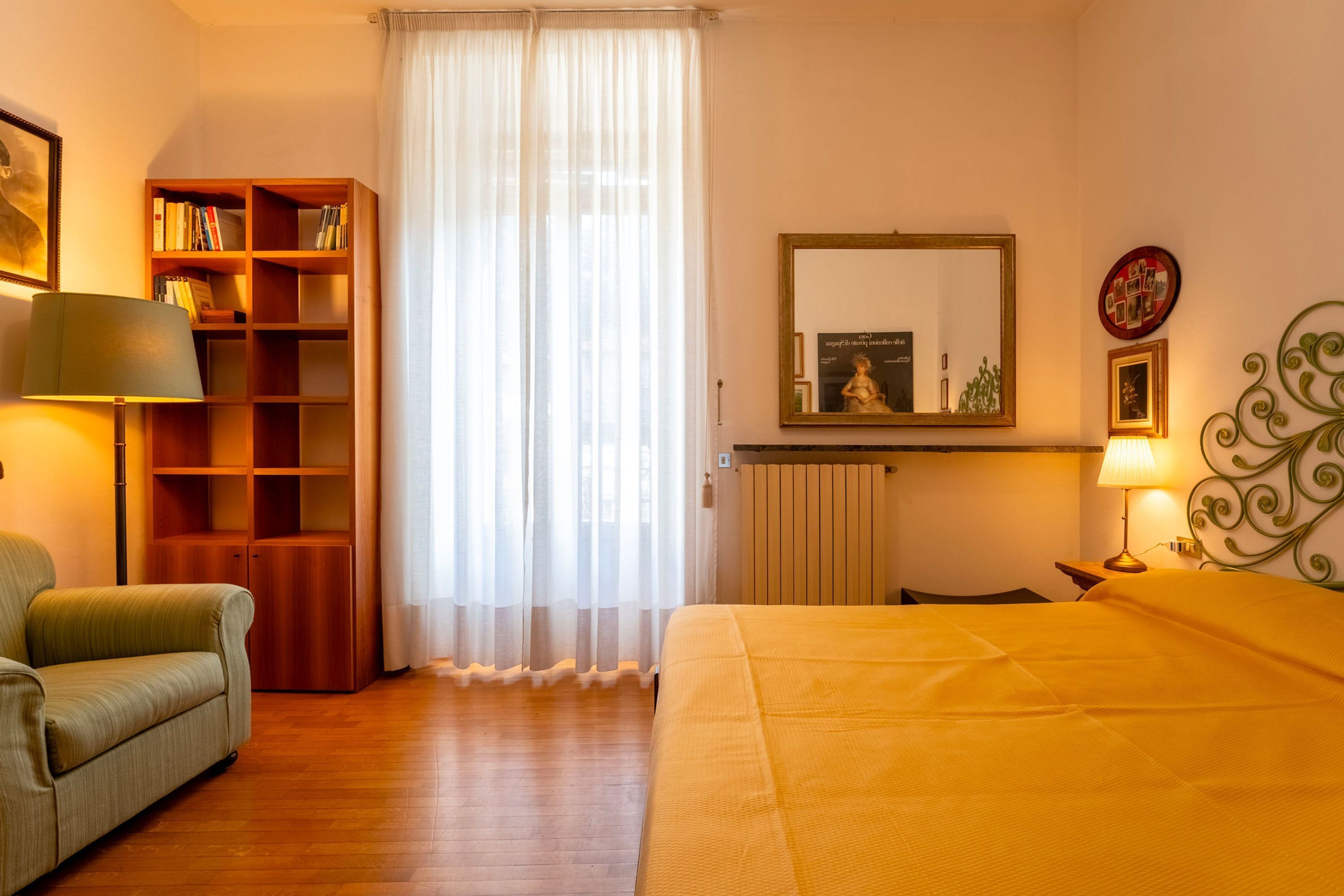 A hotel room with a bed, a sofa, a bookshelf, a window with sheer curtains, a radiator, and a wall-mounted mirror.