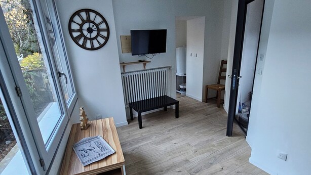 A modern living room with a wooden floor, a TV mounted on the wall, a radiator, and a window with a view of greenery. - Superb cosy studio for 2 people - Luchon center (Bagnères-de-Luchon)