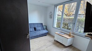 A modern living room with a blue sofa, a wooden coffee table, and a large window offering a view of trees and a fence. - Superb cosy studio for 2 people - Luchon center (Bagnères-de-Luchon)