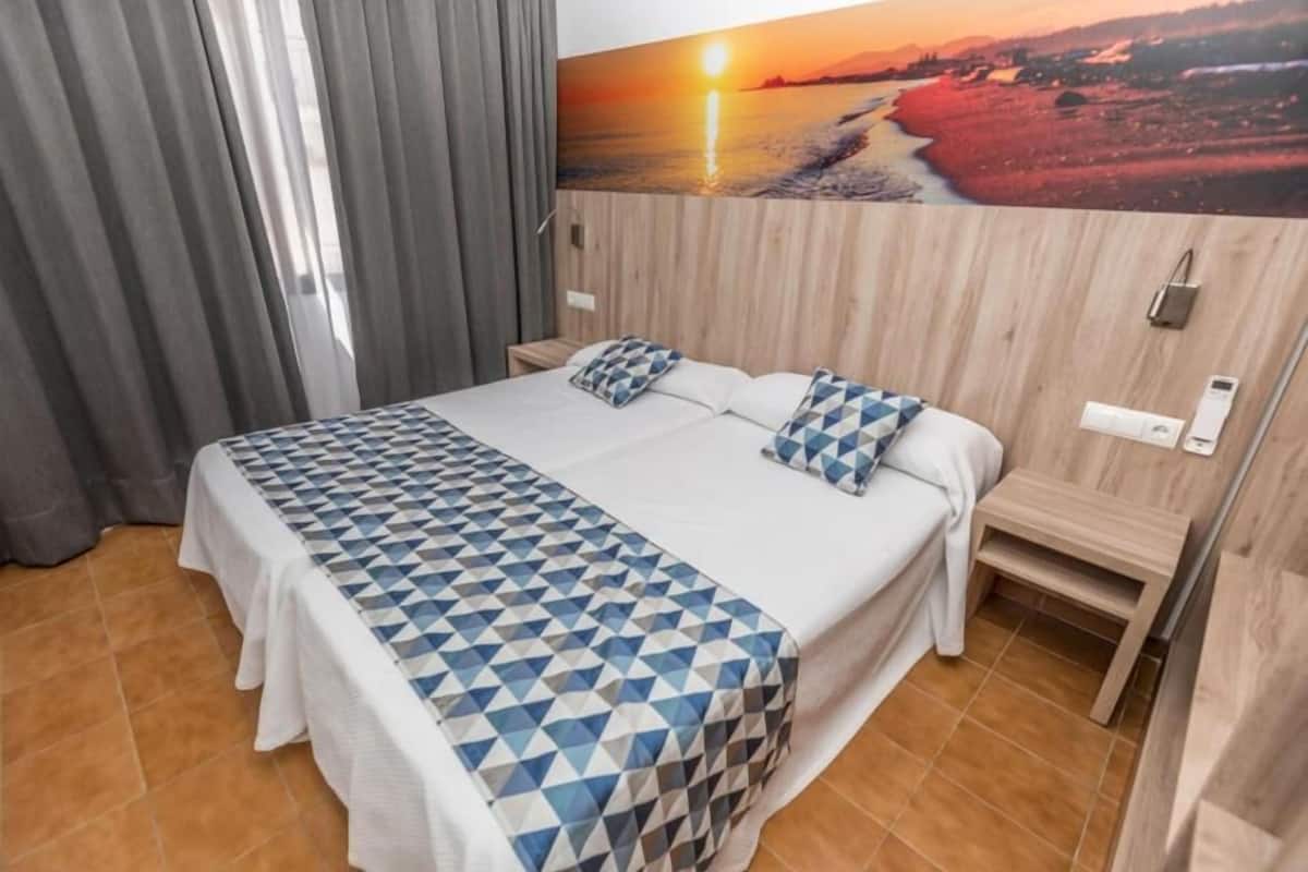 Ein Hotelzimmer mit einem großen Bett, einem Nachttisch, einem hölzernen Kopfteil, einem Gemälde mit Sonnenuntergang über einem Strand und einem Fenster mit Vorhängen.