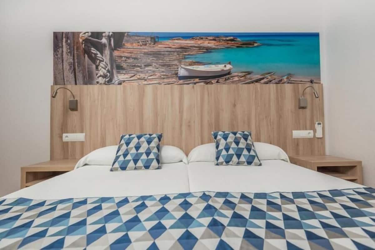 Ein Hotelzimmer mit einem Bett, zwei Kissen, einem hölzernen Kopfteil, einem Bild einer Küstenlandschaft und einem Nachttisch.