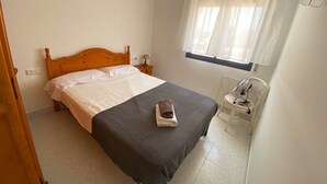 Un lit bien fait, avec une tête de lit en bois, une table de chevet avec une lampe, une fenêtre ornée de rideaux légers et un radiateur blanc.