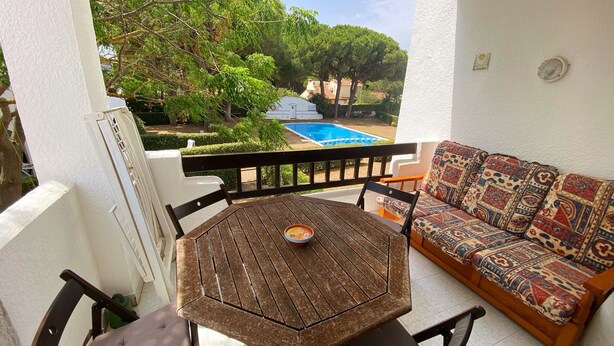 Un balcon avec une table et des chaises en bois, un canapé agrémenté de coussins à motifs et une vue sur une piscine et la verdure.
