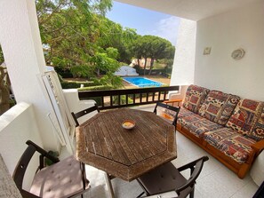 Un balcon avec une table et des chaises en bois, un canapé agrémenté de coussins à motifs et une vue sur une piscine et la verdure.