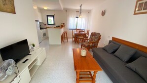 Un salon comprenant un canapé, une table basse, un téléviseur et un ventilateur. Il y a également un coin cuisine avec une table à manger et des chaises.
