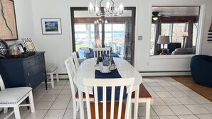 A dining area with a white table, blue chairs, and a chandelier. There is a view of the outdoors through large windows.