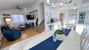 A modern living room with a blue sofa, a dining table with a blue runner, and a kitchen area with white cabinets and a refrigerator.