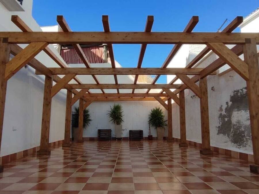 Un espacio al aire libre cubierto con una pérgola de madera, suelo de baldosas y plantas en macetas.